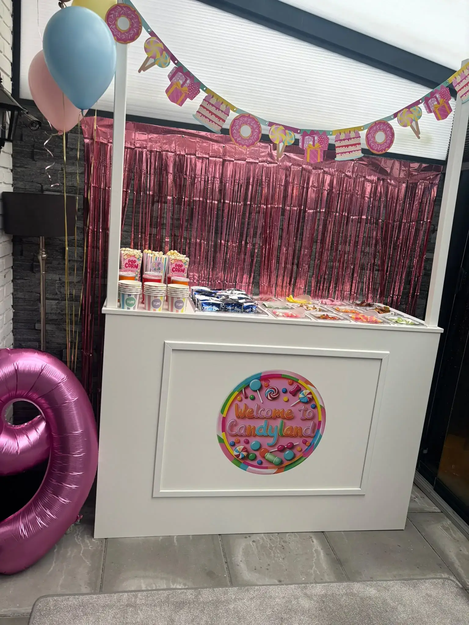Witte candy bar kraam met roze folie, een feestslinger, popcornbakjes en snoepbakken